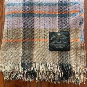 K&ouml;nnte beinhalten: Eine gefaltete Wolldecke mit einem Karomuster in den Farbt&ouml;nen Lavendel, Braun, Orange und Blau. Die Decke hat einen Fransenrand und ein schwarzes Etikett mit der Aufschrift "EARLYS OF WITNEY" und "All Wool Throw."