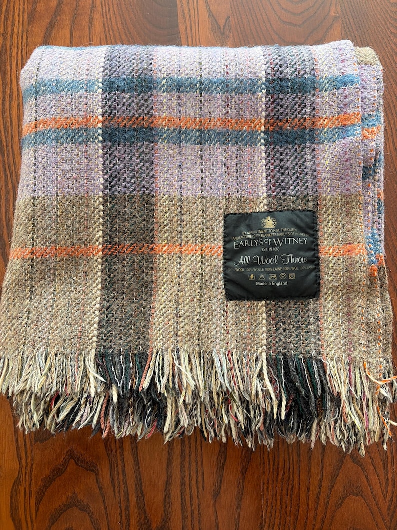 K&ouml;nnte beinhalten: Eine gefaltete Wolldecke mit einem Karomuster in den Farbt&ouml;nen Lavendel, Braun, Orange und Blau. Die Decke hat einen Fransenrand und ein schwarzes Etikett mit der Aufschrift "EARLYS OF WITNEY" und "All Wool Throw."
