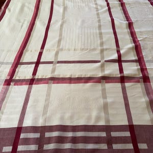 May include: A rectangular tablecloth featuring a cream-colored base with a prominent plaid pattern in shades of deep red and muted beige. The fabric displays a subtle, woven damask-style texture throughout, with a wide, solid red border at the end.