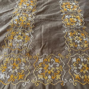 May include: A brown textile with intricate embroidery in yellow and white. The embroidered design features floral and geometric patterns arranged in a symmetrical border and central design. The fabric has a slightly wrinkled texture.