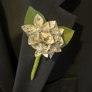 May include: A boutonniere made of white paper flowers with musical notes printed on them. The flowers are accented with small white pearls and green leaves.