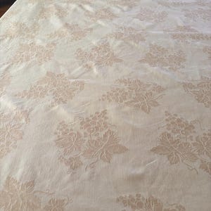 May include: A cream-colored tablecloth featuring a repeating jacquard pattern of grape clusters and vine leaves. The fabric has a subtle sheen, and the design is woven directly into the textile, creating a textured, tonal appearance across the surface.
