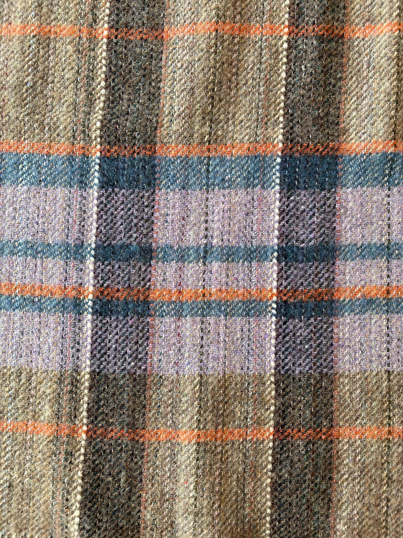 K&ouml;nnte beinhalten: Nahaufnahme eines gewebten Stoffes mit einem Karomuster. Das Design zeigt vertikale und horizontale Streifen in Braun-, Grau-, Blau-, Orange- und Lavendelt&ouml;nen. Der Stoff hat ein strukturiertes Aussehen.