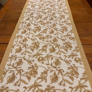 May include: A rectangular table runner featuring a white fabric base with a repeating tan-colored botanical print of pomegranate fruits, leaves, and winding vines. The runner is finished with a solid tan fabric border along both long edges.
