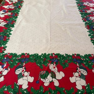 May include: A rectangular tablecloth featuring a white center with small red dots, surrounded by a wide red border. The border displays a pattern of green holly leaves, red berries, blue bells, candy canes, and white teddy bears wearing hats.