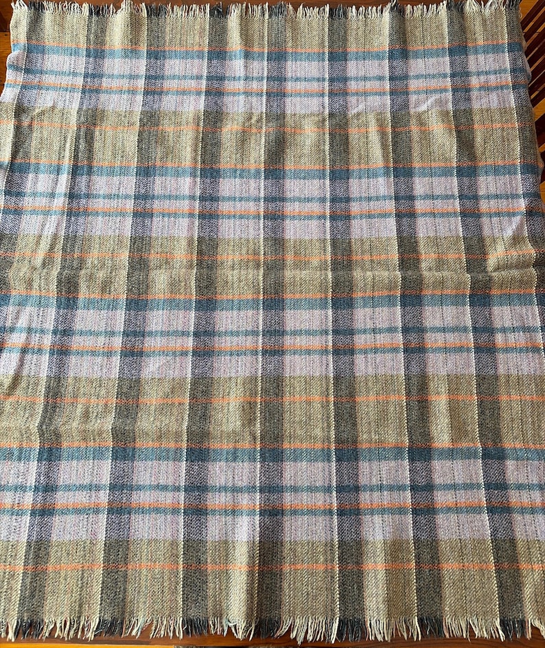 K&ouml;nnte beinhalten: Eine gewebte Decke mit einem Karomuster in Gr&uuml;n, Blau, Orange und Grau. Die Decke hat einen Fransenrand und ein klassisches Karomuster. Die Farben sind ged&auml;mpft, was ihr einen Vintage- oder rustikalen Look verleiht.