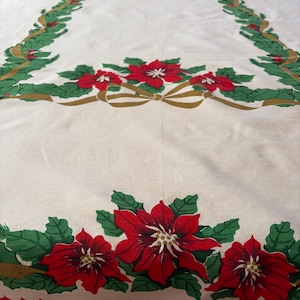 May include: A rectangular off-white tablecloth featuring a printed border of green holly leaves, red poinsettia flowers, and gold ribbons. The design includes a central rectangular garland motif and a scalloped red edge at the bottom.