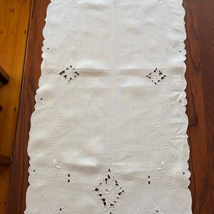 May include: White rectangular table runner with scalloped edges. The runner features embroidered floral and geometric designs, including diamond shapes and dotted lines. The fabric is a light, neutral color, suitable for various home decor styles.
