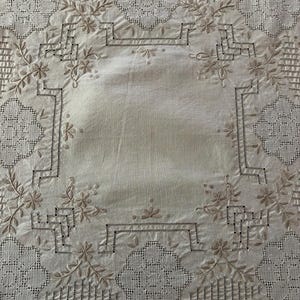 May include: A square off-white textile featuring intricate beige embroidery. The design includes floral motifs, geometric openwork patterns, and delicate needlework borders arranged in a symmetrical layout across the fabric surface.
