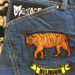 May include: Blue denim jeans with a black patch that says "Versace" and two embroidered patches. One patch features an orange tiger and the other a yellow banner with the word "DELIRIUM" in black.