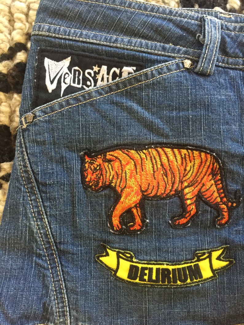 May include: Blue denim jeans with a black patch that says "Versace" and two embroidered patches. One patch features an orange tiger and the other a yellow banner with the word "DELIRIUM" in black.