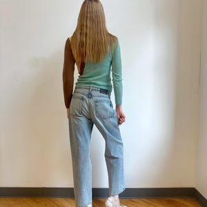 May include: A woman wearing a color-block sweater in brown, yellow, and mint green, paired with light-wash denim jeans. The outfit is completed with white, sculptural heels. The model is standing in front of a white wall and wooden floor.