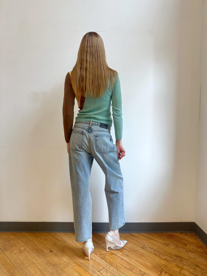 May include: A woman wearing a color-block sweater in brown, yellow, and mint green, paired with light-wash denim jeans. The outfit is completed with white, sculptural heels. The model is standing in front of a white wall and wooden floor.
