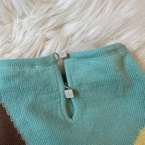 May include: Close-up of a knitted top with a keyhole neckline and two square, off-white buttons. The top features a color block design with shades of turquoise, brown, and yellow. The fabric has a textured appearance.