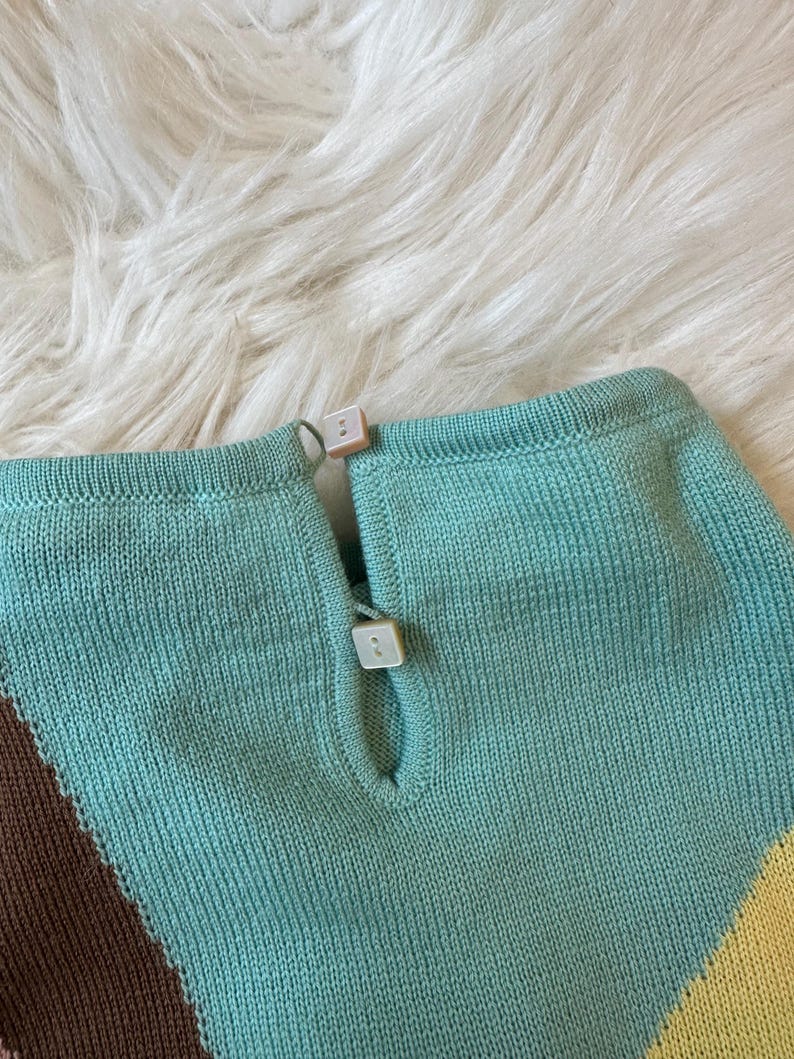 May include: Close-up of a knitted top with a keyhole neckline and two square, off-white buttons. The top features a color block design with shades of turquoise, brown, and yellow. The fabric has a textured appearance.