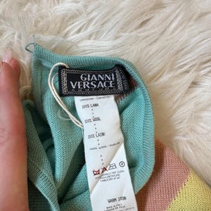 May include: A close-up of a Gianni Versace label attached to a teal, pink, and yellow striped wool garment. The label features the Gianni Versace logo and care instructions. The garment is made in Italy.