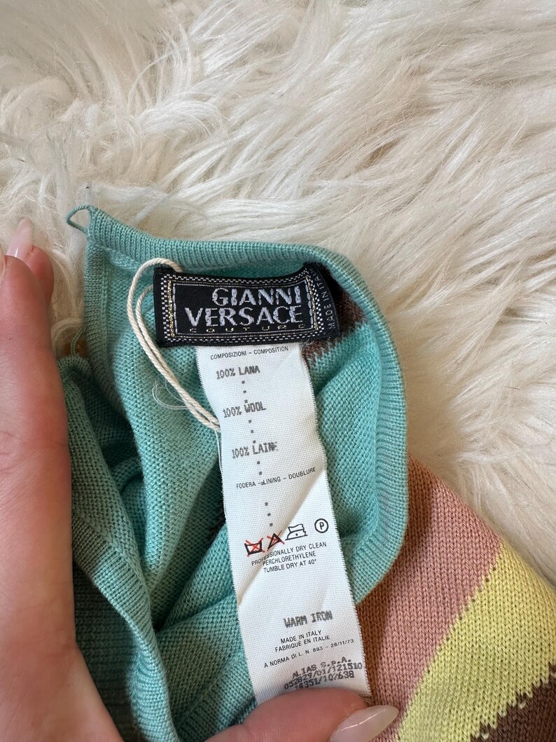 May include: A close-up of a Gianni Versace label attached to a teal, pink, and yellow striped wool garment. The label features the Gianni Versace logo and care instructions. The garment is made in Italy.