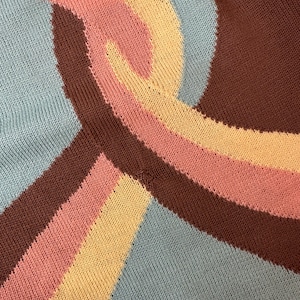 May include: Close-up of a knitted textile with a repeating wave pattern. The design features a combination of colors, including light blue, brown, pink, and yellow. The fabric has a textured appearance, with visible knit stitches.