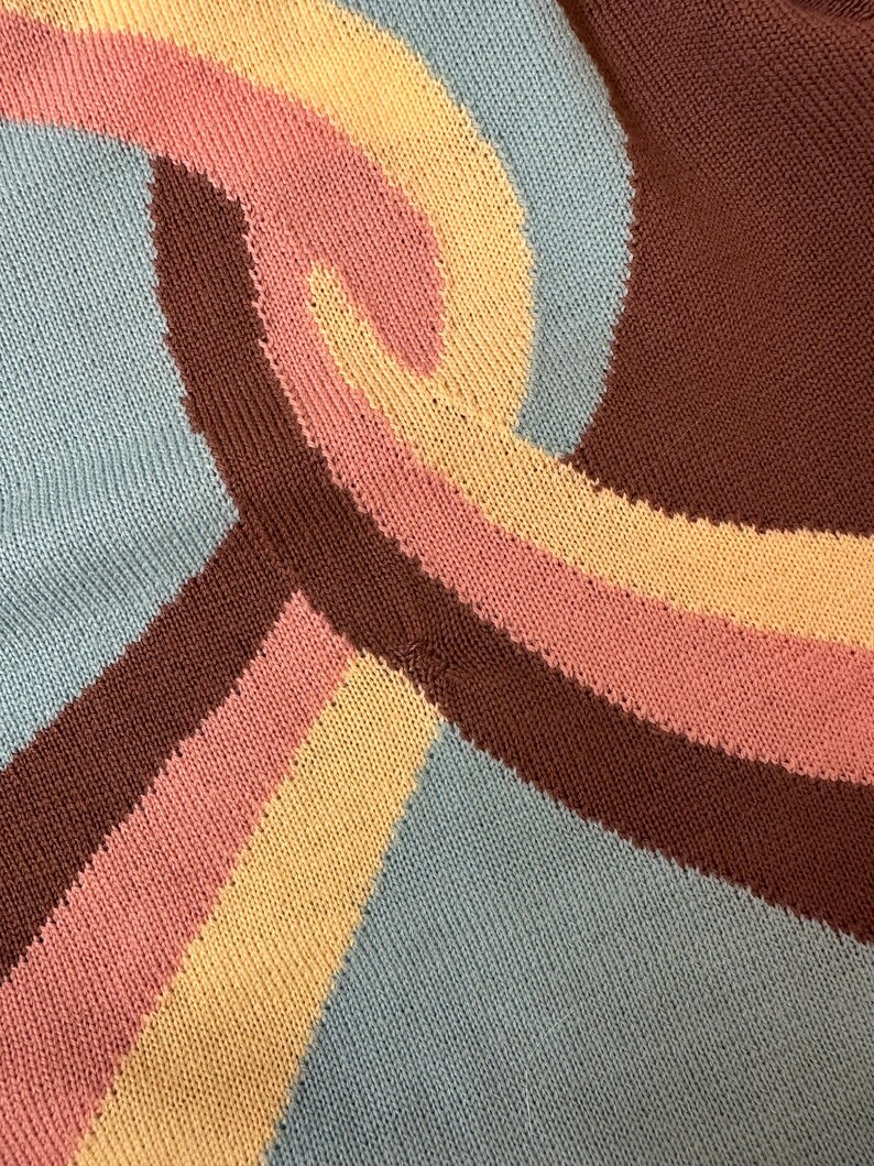 May include: Close-up of a knitted textile with a repeating wave pattern. The design features a combination of colors, including light blue, brown, pink, and yellow. The fabric has a textured appearance, with visible knit stitches.