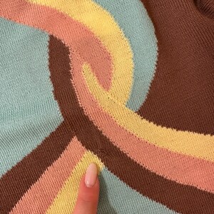 May include: Close-up of a knitted fabric with a retro design. The pattern features curved stripes in shades of brown, light blue, yellow, and peach. The fabric has a textured appearance, suggesting a sweater or similar garment.