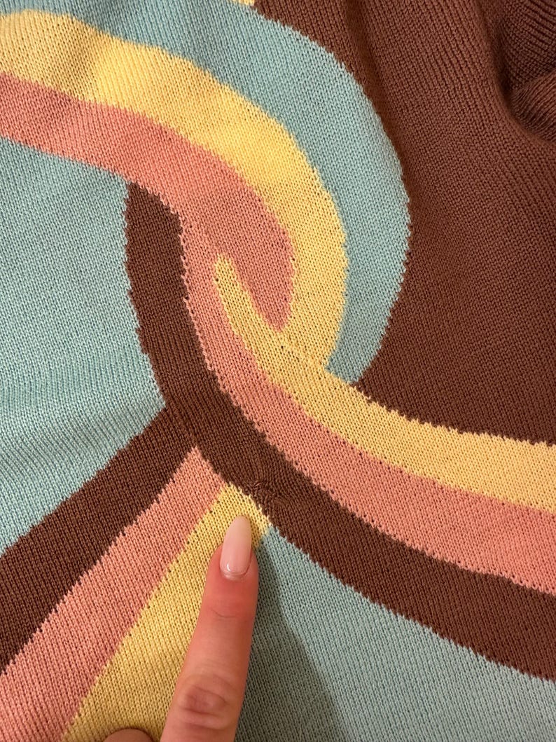 May include: Close-up of a knitted fabric with a retro design. The pattern features curved stripes in shades of brown, light blue, yellow, and peach. The fabric has a textured appearance, suggesting a sweater or similar garment.