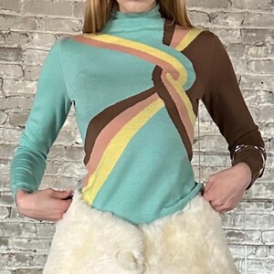 May include: A long-sleeved turtleneck sweater with a unique design. The sweater features a turquoise body with brown, yellow, and pink stripes. One sleeve is brown. The sweater has a retro, 70s-inspired aesthetic.