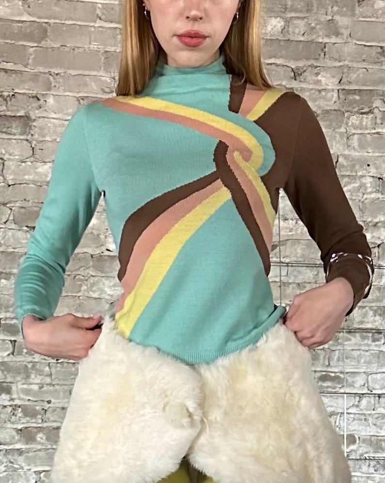 May include: A long-sleeved turtleneck sweater with a unique design. The sweater features a turquoise body with brown, yellow, and pink stripes. One sleeve is brown. The sweater has a retro, 70s-inspired aesthetic.