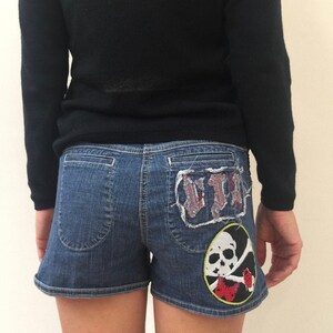 May include: Blue denim shorts with a skull and crossbones patch and a red, white, and blue patch on the back pockets.