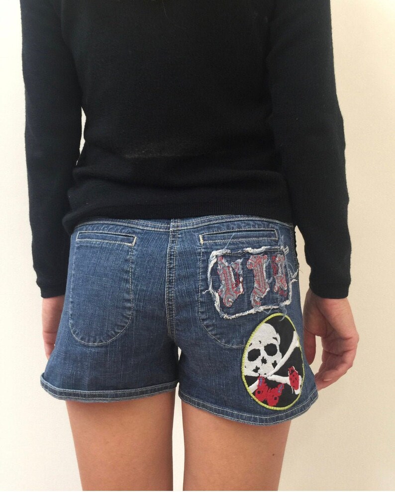 May include: Blue denim shorts with a skull and crossbones patch and a red, white, and blue patch on the back pockets.