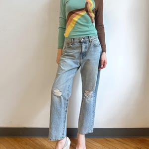 May include: A woman wearing a teal turtleneck sweater with a colorful abstract design, light blue ripped jeans, and white heels. The sweater features brown, yellow, and pink accents. The jeans have distressed details at the knees. The shoes have a pointed toe.