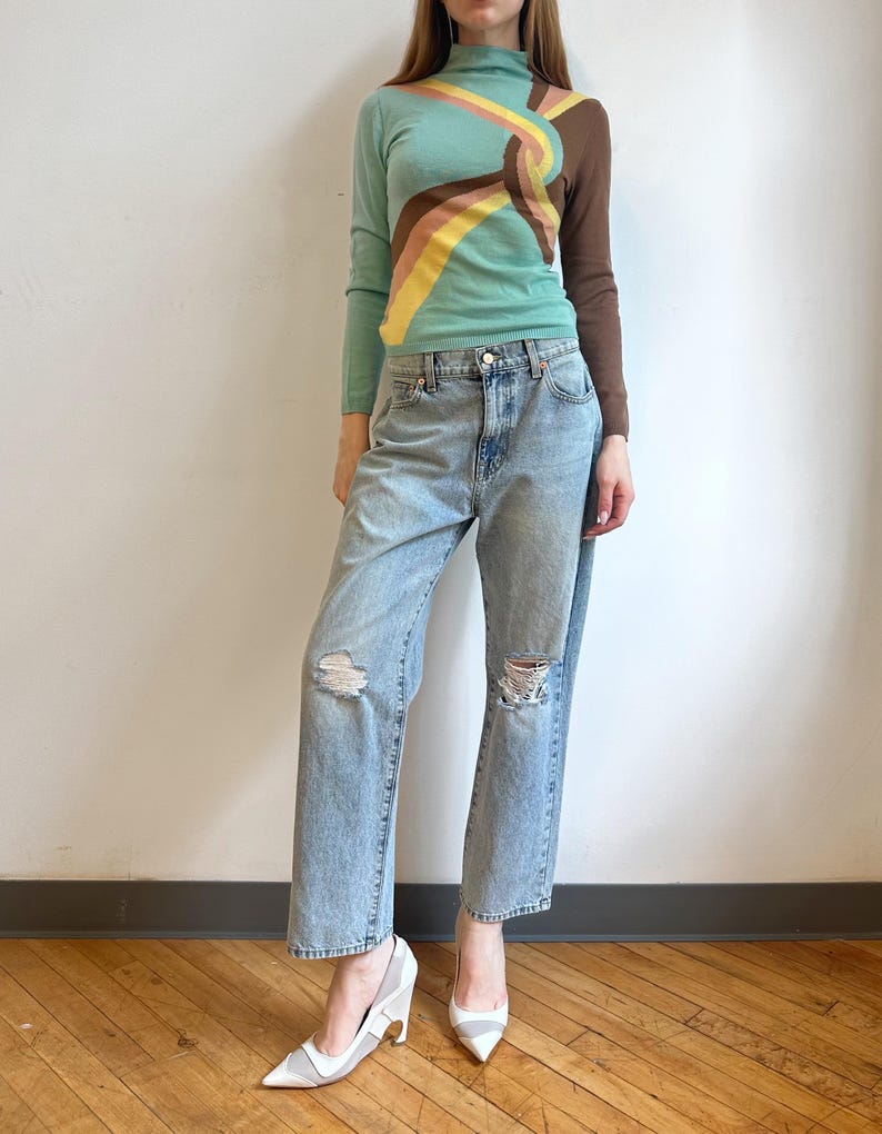 May include: A woman wearing a teal turtleneck sweater with a colorful abstract design, light blue ripped jeans, and white heels. The sweater features brown, yellow, and pink accents. The jeans have distressed details at the knees. The shoes have a pointed toe.
