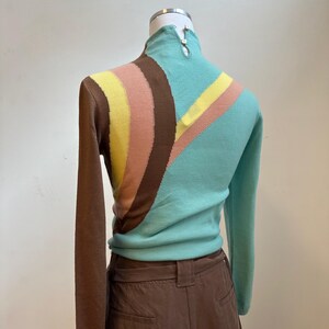 May include: A vintage-style sweater with a high collar and long sleeves. The sweater features a color block design with brown, yellow, pink, and teal stripes. The sweater is displayed on a mannequin.
