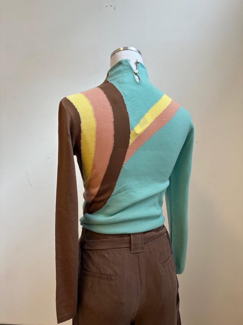 May include: A vintage-style sweater with a high collar and long sleeves. The sweater features a color block design with brown, yellow, pink, and teal stripes. The sweater is displayed on a mannequin.