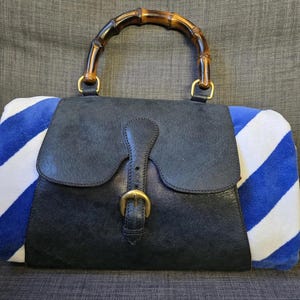 May include: A black leather handbag with a bamboo handle and gold-tone hardware. The sides are made of white and blue striped fabric. The bag has a flap closure with a buckle.