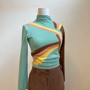 May include: A long-sleeved, mock turtleneck sweater in a light teal color with a geometric design. The design includes stripes of brown, yellow, and pink. The sweater is paired with brown pants.