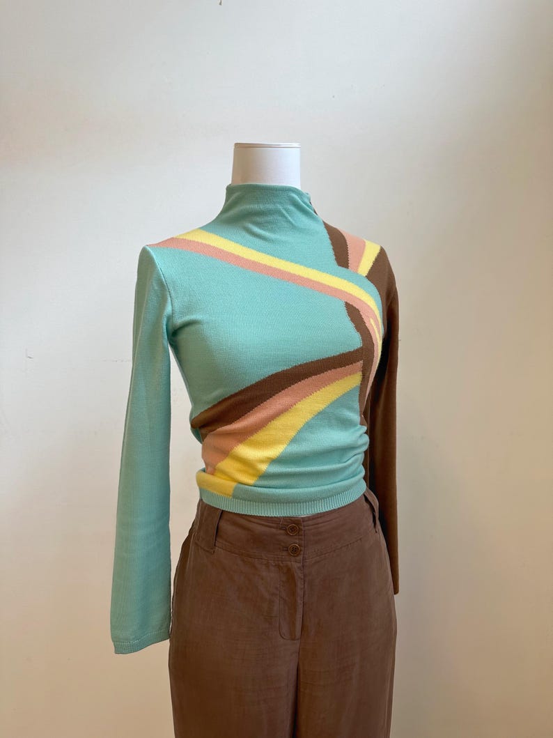 May include: A long-sleeved, mock turtleneck sweater in a light teal color with a geometric design. The design includes stripes of brown, yellow, and pink. The sweater is paired with brown pants.