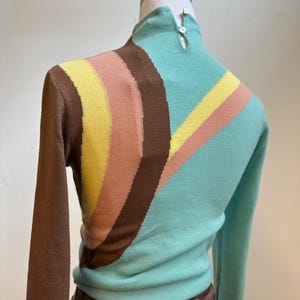 May include: A vintage-style sweater with a high neck and long sleeves. The sweater features a color block design with brown, yellow, peach, and teal stripes across the chest and sleeves. The back of the neck has two small buttons.