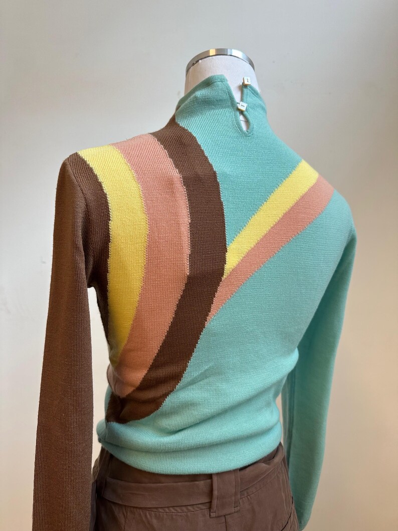May include: A vintage-style sweater with a high neck and long sleeves. The sweater features a color block design with brown, yellow, peach, and teal stripes across the chest and sleeves. The back of the neck has two small buttons.