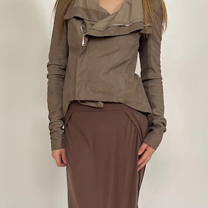May include: A brown jacket with an asymmetrical zipper and a draped collar is paired with a long brown skirt. The jacket has long sleeves and a tailored fit. The skirt is a solid color and has a flowing silhouette.