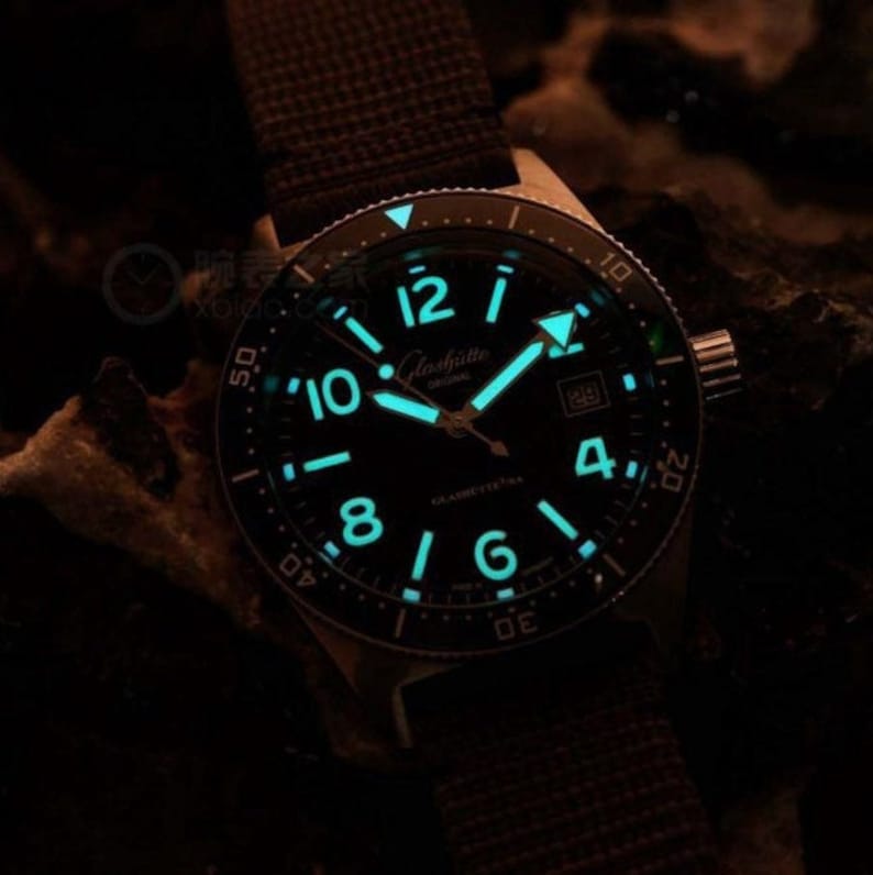 May include: A black watch with a luminous dial and hands. The watch has a black bezel with white markings and a black leather strap.