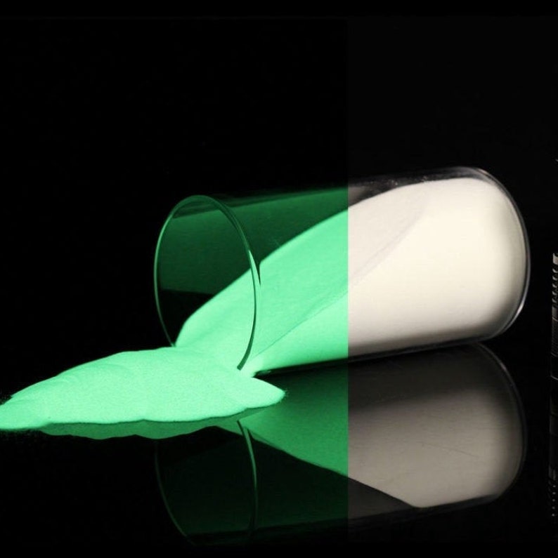 May include: A glass jar filled with glowing green sand, spilling out onto a black surface. The sand glows brightly in the dark.