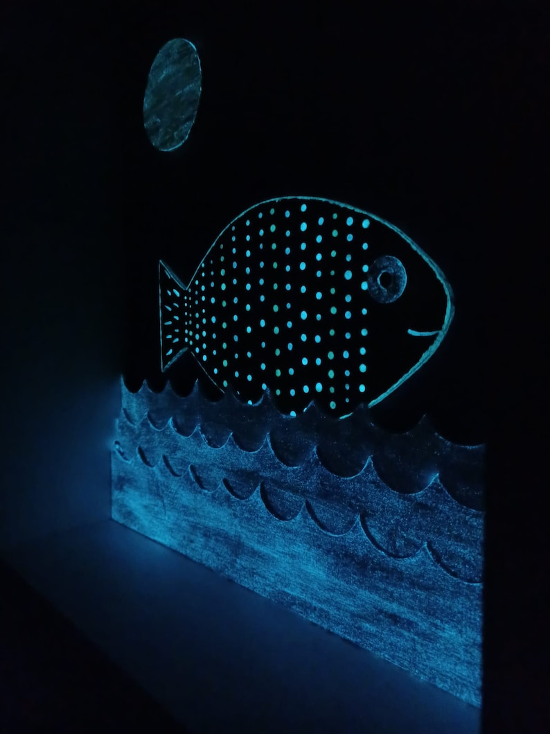 May include: A black and white fish with a blue glow, swimming in a blue ocean with a white moon in the background.