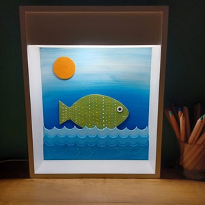 May include: A framed artwork depicting a green fish with white spots swimming in blue water with white waves. A yellow sun is in the upper left corner of the image.