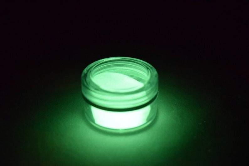May include: A small, round jar filled with a bright green glow-in-the-dark powder. The jar is made of clear glass and has a black lid.