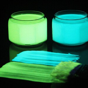 May include: Two glass jars of glow-in-the-dark paint, one green and one blue, with a paintbrush and streaks of paint on a black surface.