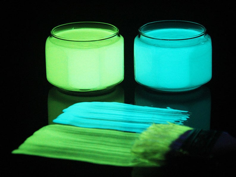 May include: Two glass jars of glow-in-the-dark paint, one green and one blue, with a paintbrush and streaks of paint on a black surface.