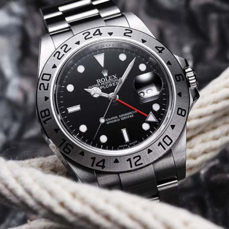 May include: A silver Rolex Explorer II watch with a black face and a red second hand. The watch has a 24-hour bezel and a date window at 3 o'clock. The watch is on a silver bracelet.