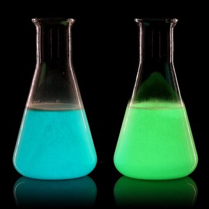 May include: Two glass beakers filled with glowing liquid. The beaker on the left contains a bright blue liquid, while the beaker on the right contains a bright green liquid.