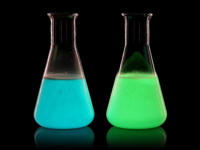 May include: Two glass beakers filled with glowing liquid. The beaker on the left contains a bright blue liquid, while the beaker on the right contains a bright green liquid.