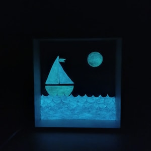 May include: A glow-in-the-dark wooden box with a sailboat and moon design. The sailboat is white with a yellow sail and is on a blue sea. The moon is yellow and is in the upper right corner of the box.